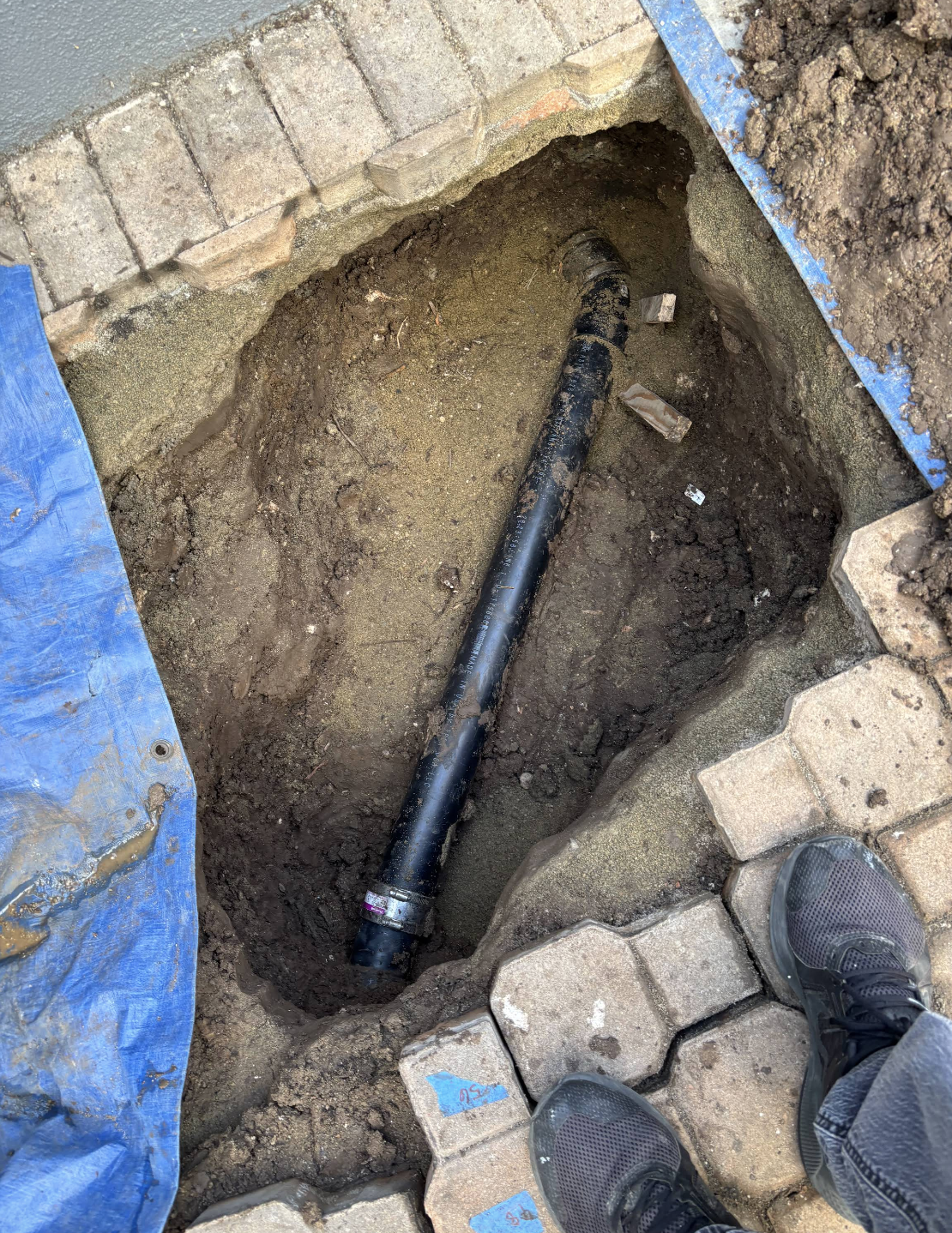 Underground black pipe installation in excavated trench with blue sheeting and concrete pavers